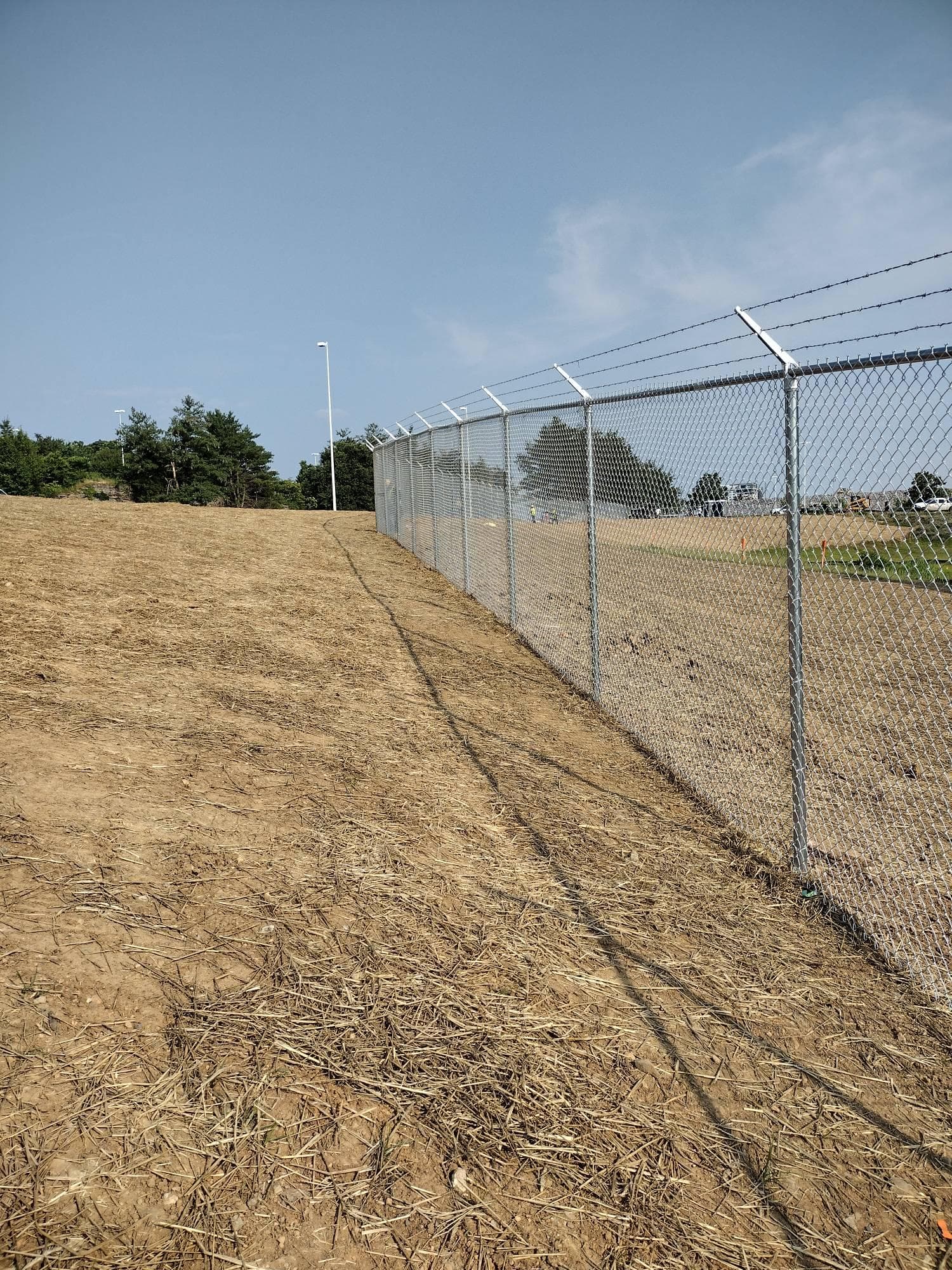 A tall galvanized chain-link security fence topped with barbed wire running along a graded commercial site