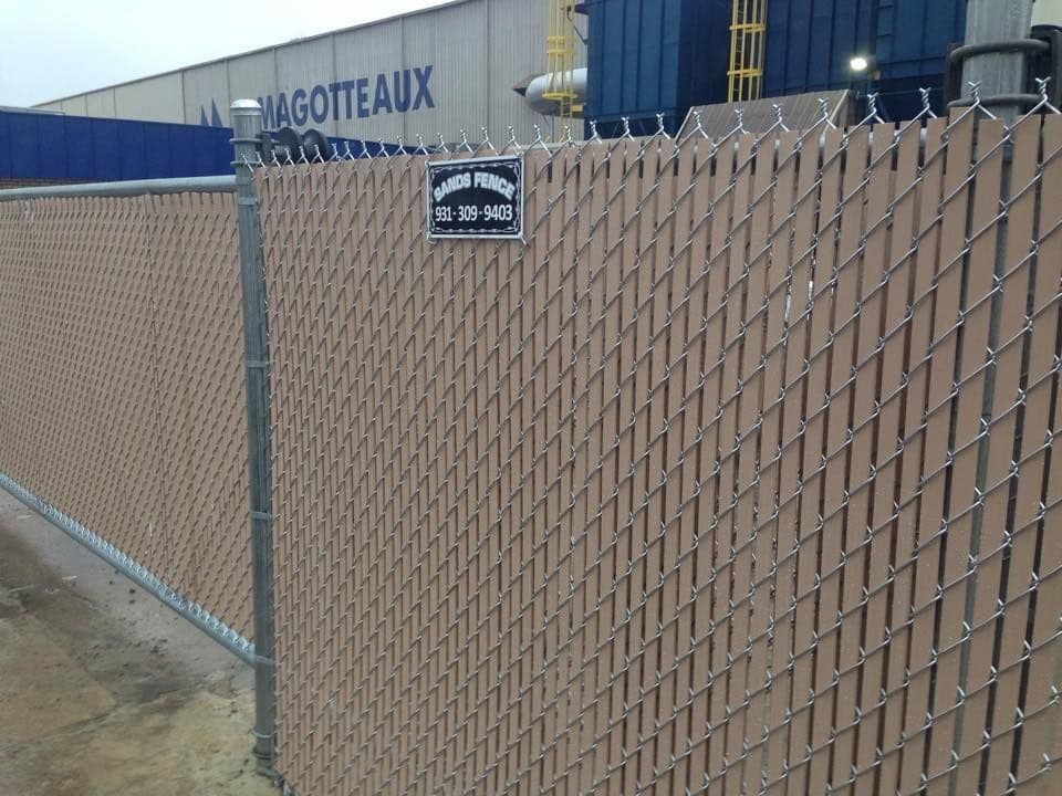A brown privacy-slat chain-link fence with a Sands Fence sign in front of an industrial building
