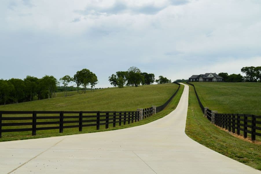 A black four-board fence with stone columns flanking a paved driveway leading to a large estate home