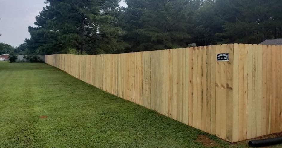 A long natural-wood privacy fence with dog-ear pickets running along a green lawn, with a Sands Fence Co. sign mounted on it