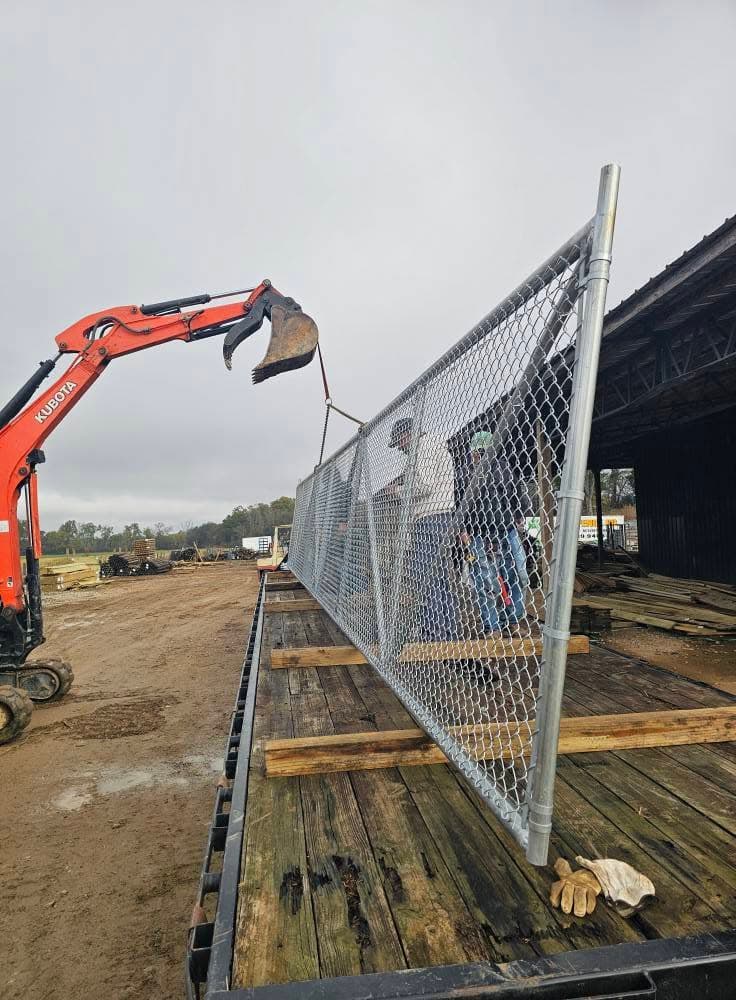 A galvanized chain-link fence panel lifted by an excavator onto a flatbed trailer at the Sands Fence Co. yard