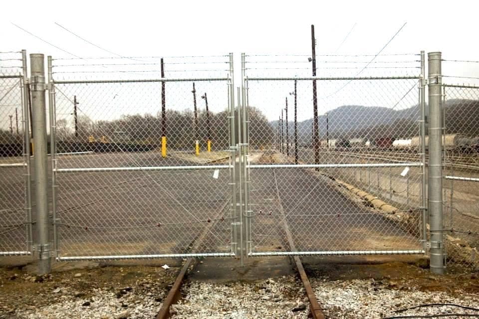 A galvanized chain-link double swing gate with barbed-wire top installed across railroad tracks at an industrial rail yard