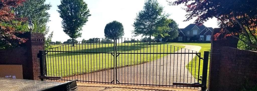 A black ornamental driveway gate with brick columns opening to an estate lawn and home