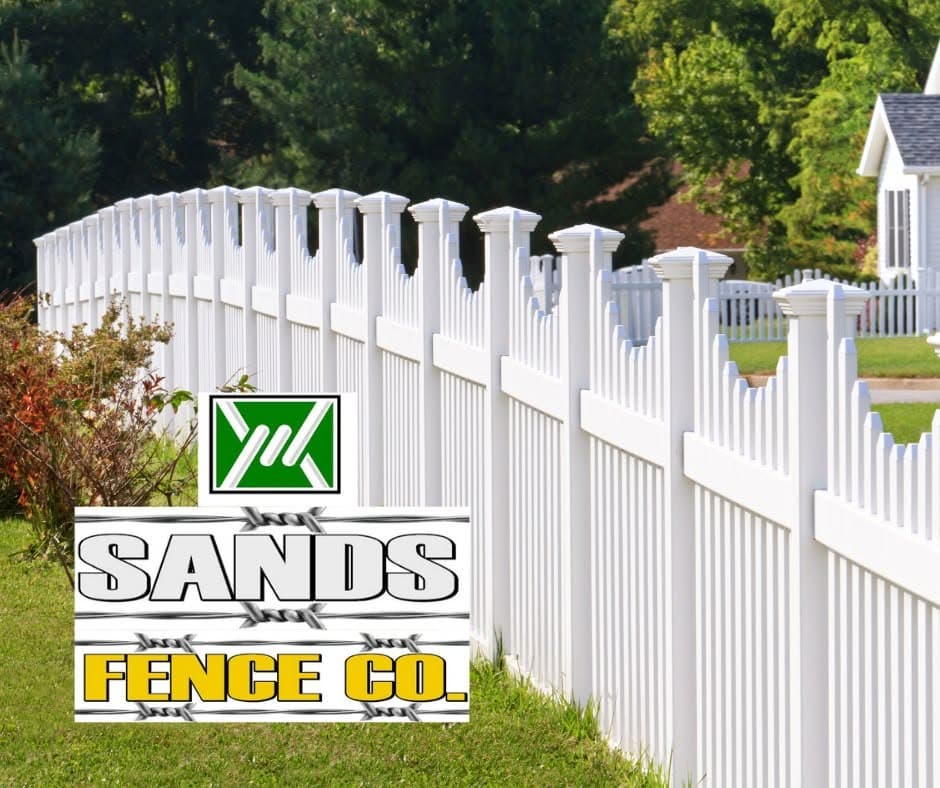 A white vinyl scalloped picket fence running along a landscaped suburban yard with a Sands Fence Co. sign