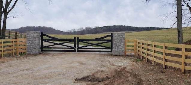Photo of double stylized black gate framed by stacked stone. The fencing for the black gate is a horizontal wood slat fence