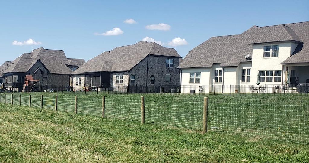 Photo of a metal fencing with wood posts that runs behind an entire subdivision row of homes. Several homes have individual property fences. The metal fence delineates the common grounds property line, separating the subdivision from the roadway